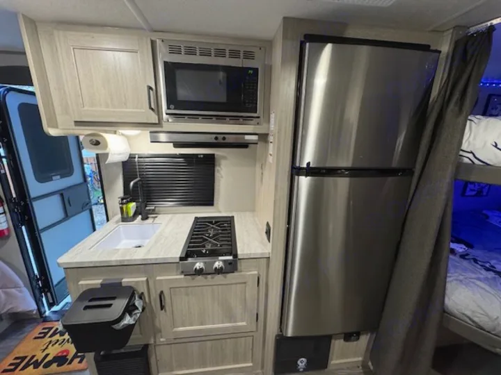 RV rental interior: compact kitchen with microwave, stove, sink, fridge. Bunk beds with privacy curtain visible right.. Forest river Aurora 2025