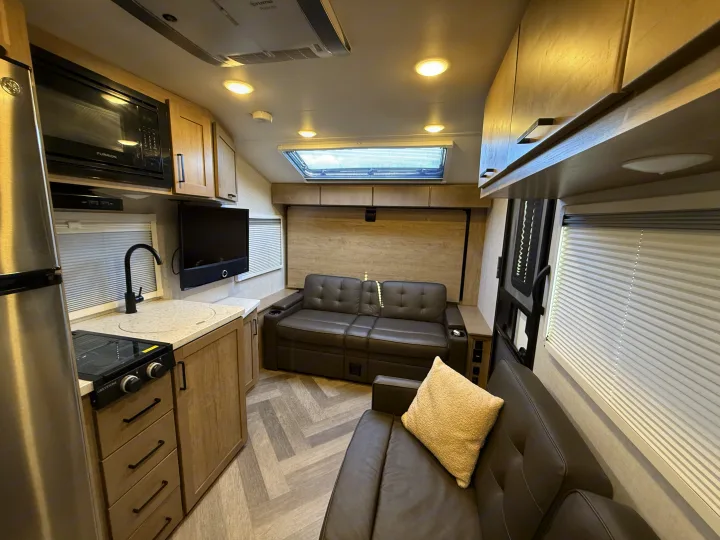 Inside this modern RV rental: a bright living area with kitchen, comfy sofas, TV, and skylight.. Ember Overland 2023