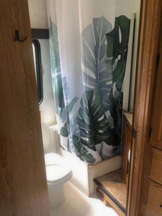 This RV rental bathroom features a compact toilet, shower with a tropical leaf curtain, and wooden cabinetry.. Ford Ford E350 1996