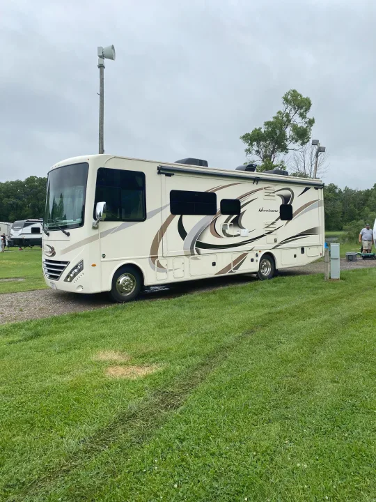 Large cream 'Hurricane' RV rental with decals parked on a grassy site at a peaceful campground. Ideal for outdoor living.. Thor Hurricane 2018