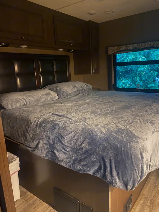 Cozy RV rental bedroom with a large bed, plush gray bedding, dark headboard, overhead cabinets, and a window showing trees outside.. Thor Hurricane 2018