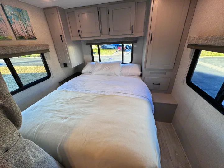 Cozy RV rental bedroom featuring a bed with white linens, overhead cabinets, and windows offering views outside, perfect for travel.. Forest River Coachmen Freelander 2026