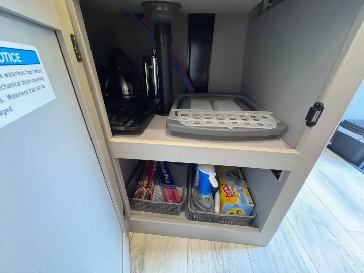 Compact under-sink storage in an rv rental, showing plumbing, a collapsible drying rack, cleaning spray, foil, and trash bags.. Forest River Coachmen Freelander 2026