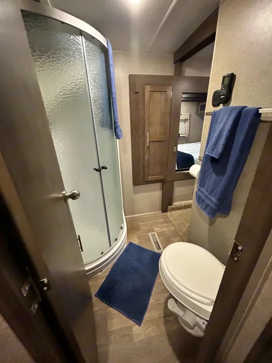 This compact RV rental bathroom features a curved shower, toilet, and mirror reflecting the bedroom area.. Forest River Flagstaff Super Lite 2019