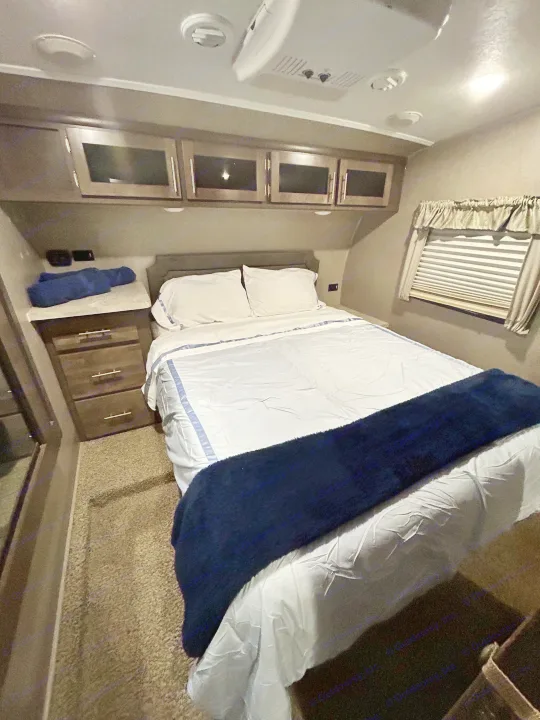 This RV rental's cozy bedroom features a bed with white and blue linens, overhead storage, and a window.. Forest River Flagstaff Super Lite 2019