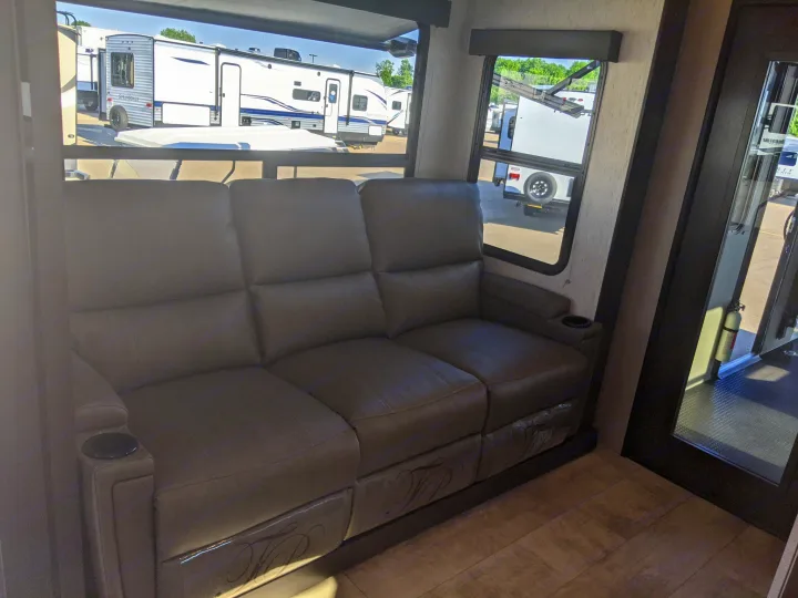 Spacious RV rental interior: a brown, comfortable three-seat reclining sofa, large windows with views of other RVs, and a living area.. Grand Design Momentum Toy Hauler 2022