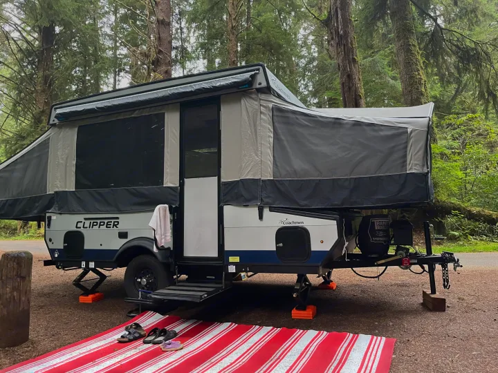 A cozy Coachmen Clipper pop-up RV rental, fully set up with awning and mat, rests among tall trees for camping.. Coachmen Clipper Sport 108ST 2022