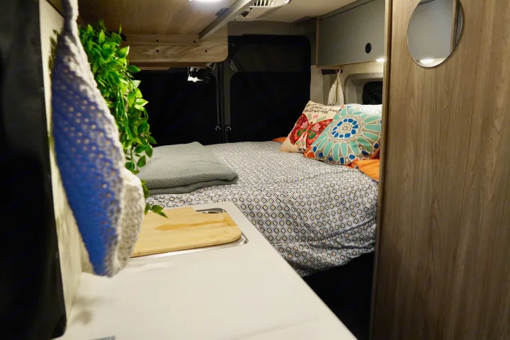 Cozy RV rental interior with a patterned bed, colorful pillows, and a compact kitchen area with a sink.. Winnebago Solis 59PX 2025