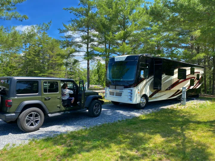 Encore RV rental set up at a scenic campsite with a green Jeep parked nearby, surrounded by tall pine trees.. Jeep Wrangler 2021