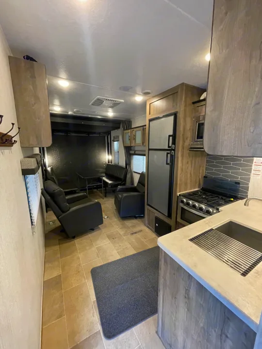 Bright rv rental interior: modern kitchen with sink, stove, and fridge, leading to a living area with black recliners and bench.. Palomino Puma 2021