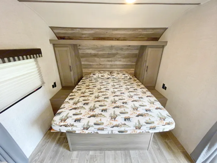 This cozy RV rental bedroom features nature-themed bedding, built-in cabinets, and a window, ideal for comfortable travel.. Gulf Stream Enlighten 2021