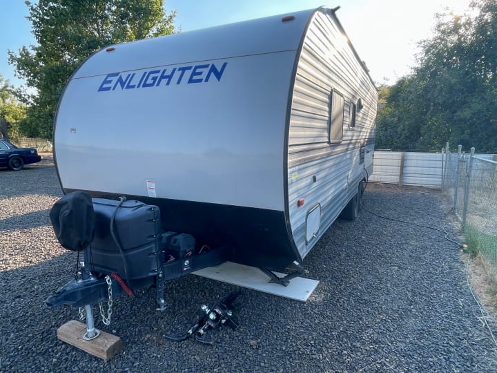 A light-colored "Enlighten" travel trailer, ready for rv rental, parked on gravel. Its hitch system is visible.. Gulf Stream Enlighten 2021