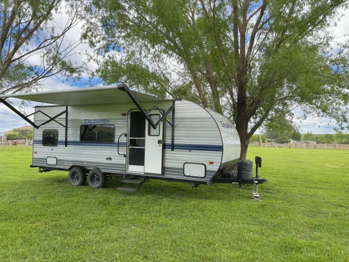 An RV rental, a gray and blue travel trailer, is parked on a green field with an extended awning under large trees.. Gulf Stream Enlighten 2021