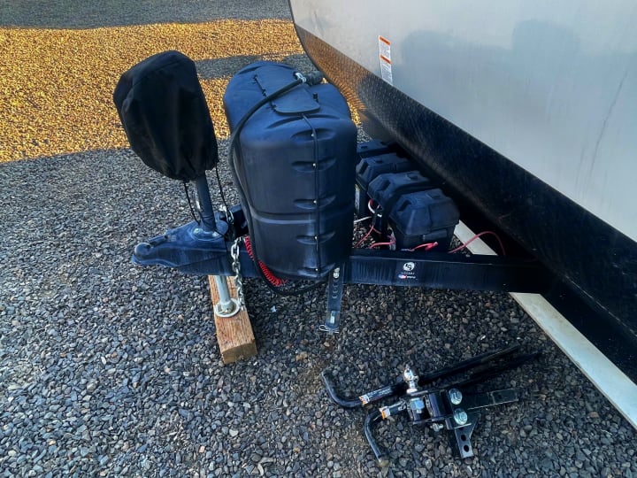 Front of an RV rental: propane tanks, batteries, hitch, and weight distribution bars rest on gravel.. Gulf Stream Enlighten 2021