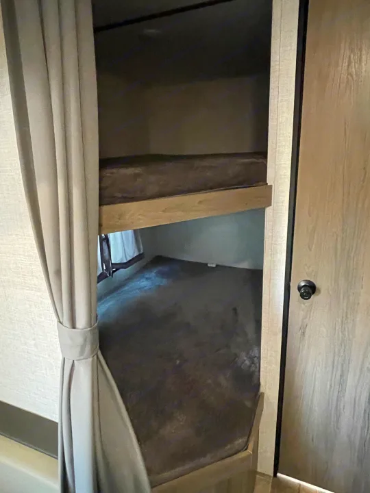Inside an RV rental, view of stacked bunk beds with dark mattresses and a tied-back curtain, for comfortable sleeping.. Gulf Stream Enlighten 2021