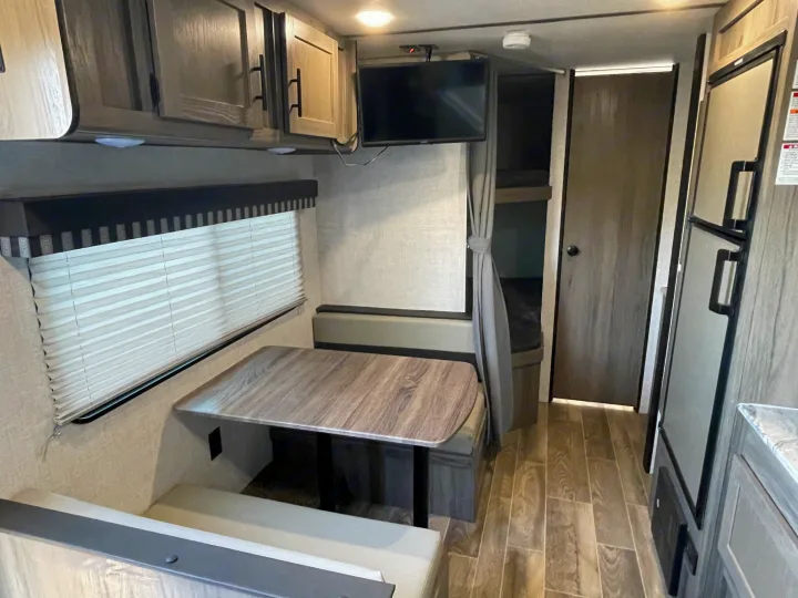 Interior of an RV rental showing a wood-top dinette, mounted TV, and bunk beds with a privacy curtain. Fridge on right.. Gulf Stream Enlighten 2021