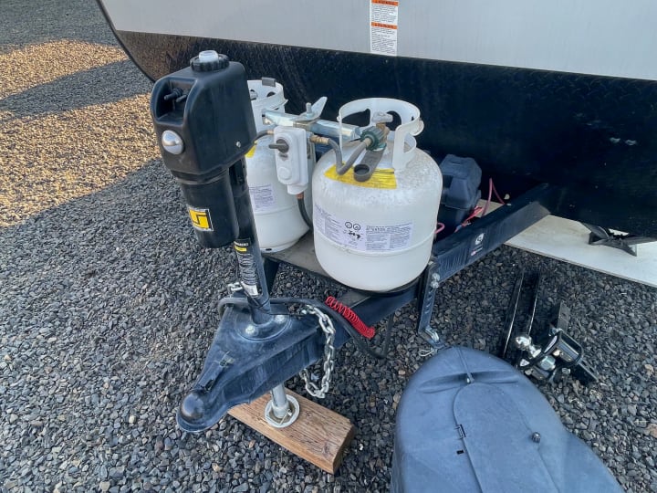 Close-up of an RV rental's front hitch with dual propane tanks and electric tongue jack on gravel.. Gulf Stream Enlighten 2021