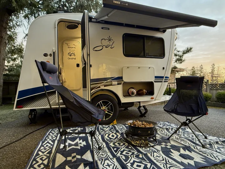 White RV rental with awning, two camp chairs, and a fire pit on a patterned rug, set up for a cozy camping trip.. InTech RV SOL 2021