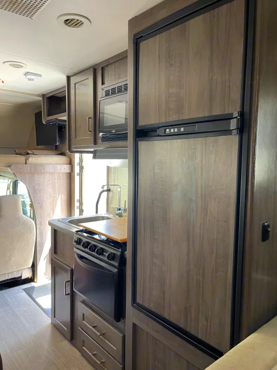 RV rental interior: A compact kitchen area featuring a dark wood-grained refrigerator, microwave, stove, and sink, with light flooring.. Winnebago Minnie Winnie 2019