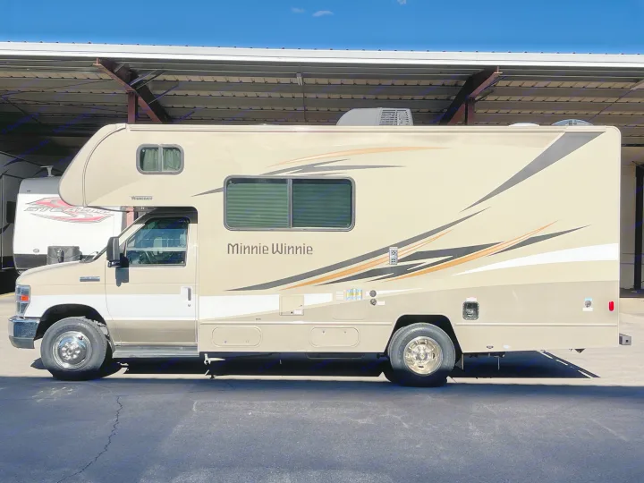 Side view of a beige Winnebago Minnie Winnie Class C RV rental, parked under a metal carport on a clear day.. Winnebago Minnie Winnie 2019