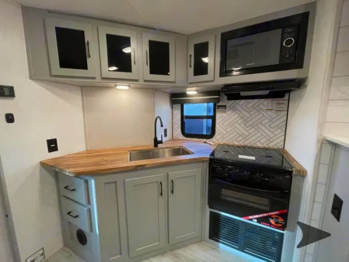 Modern RV rental kitchen with light gray cabinets, wood-look counters, sink, microwave, stove, and patterned backsplash.. Heartland RVs North Trail 2024