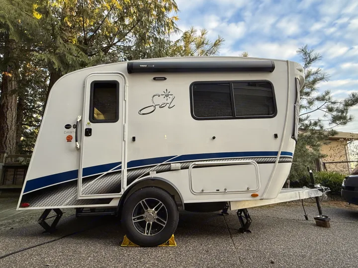 Side view of a white nuCamp Sol travel trailer RV rental, with blue stripes, parked on pavement.. InTech RV SOL 2021
