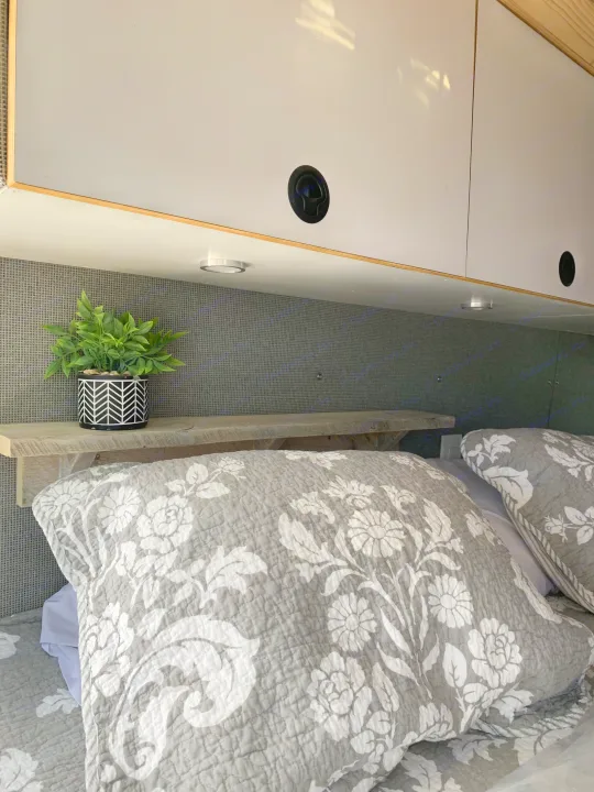 Cozy RV rental bedroom showcasing smart overhead storage, a small plant on a wood shelf, and soft floral bedding.. Ford Custom Transit 2019