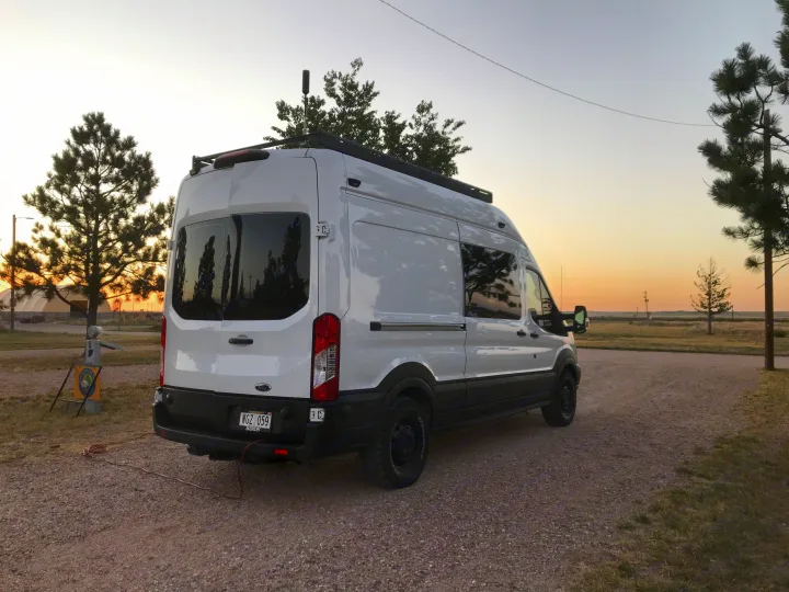 This white camper van, an RV rental, is parked on gravel at sunset, with tall trees and a campsite marker visible.. Ford Custom Transit 2019
