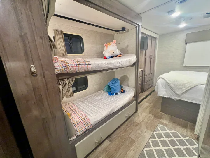 Inside an RV rental: Kids' bunk beds with windows and a cozy main bed, complete with ample storage and wood-look flooring.. Winnebago Spirit 2025
