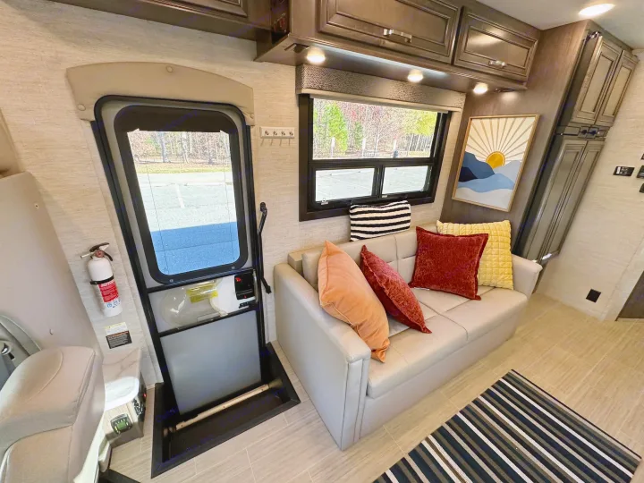 RV rental living space: a light gray sofa with colorful pillows, large window, and dark wood cabinets are visible.. Entegra Coach Esteem 2023