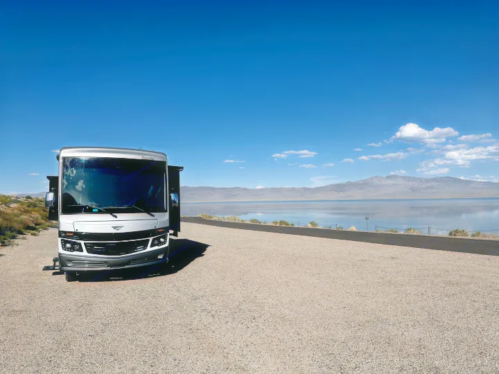Scenic view with an RV rental parked by a tranquil lake, surrounded by mountains under a clear blue sky.. Fleetwood Bounder 33C 2021