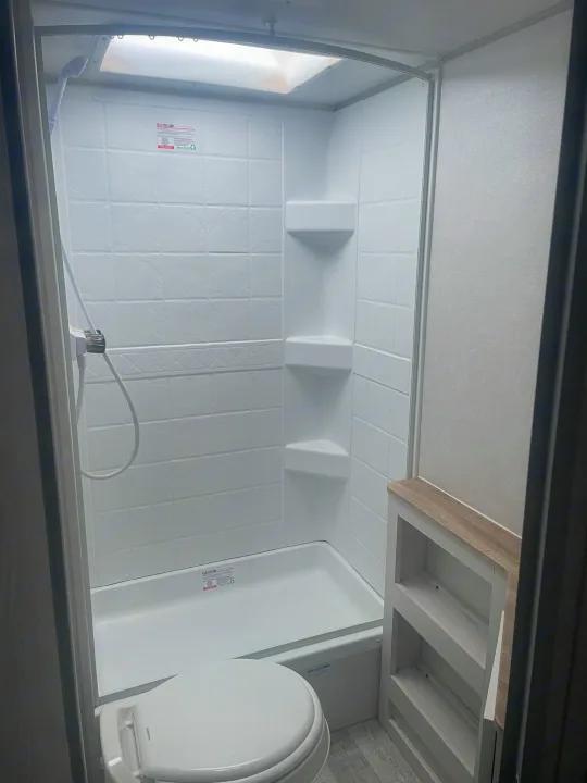 Clean RV rental bathroom: white shower, toilet, light wood-grain shelves. Bright overhead light ensures clear visibility.. No Boundaries NoBo 19.3 Essentials Only 2024