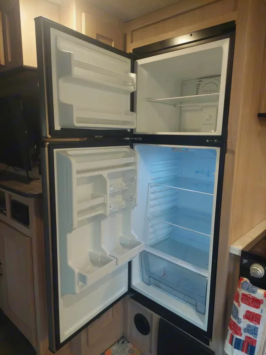 RV rental kitchen showcasing an open, empty two-door refrigerator/freezer with clear shelving and drawers, ready to be stocked.. Cruiser RV Radiance Ultra Lite 2020