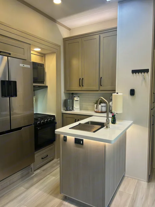 Light gray cabinets, stainless appliances, and island highlight this modern RV rental kitchen, offering comfort on the road.. Grand Design Momentum 2025