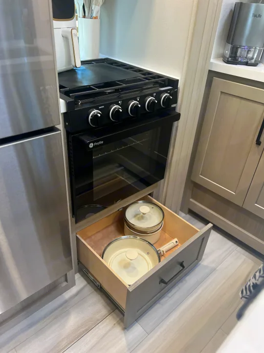 An RV rental kitchen with a black GE Profile oven/stovetop, stainless steel fridge, and an open drawer holding white pots/pans.. Grand Design Momentum 2025
