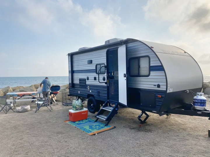 RV rental at the beach: A man grills by his camper with an ocean view, picnic table, and cozy outdoor setup.. Forest River Cherokee Wolf Pup 2020