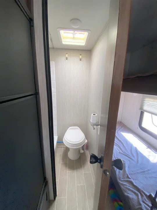 Compact RV rental bathroom with toilet, skylight, and wall hooks. An adjacent bedroom is visible through an open door.. Forest River Cherokee Wolf Pup 2020