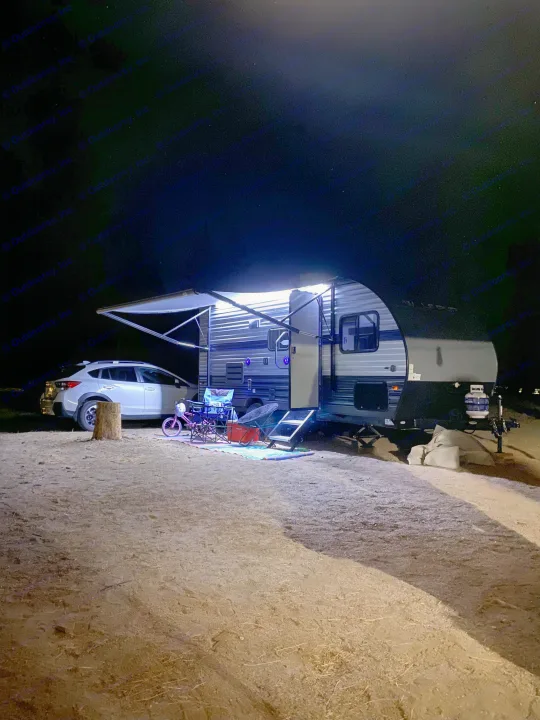Nighttime rv rental setup with bright awning lights illuminating the campsite, featuring an SUV, chairs, and a kids' bike.. Forest River Cherokee Wolf Pup 2020