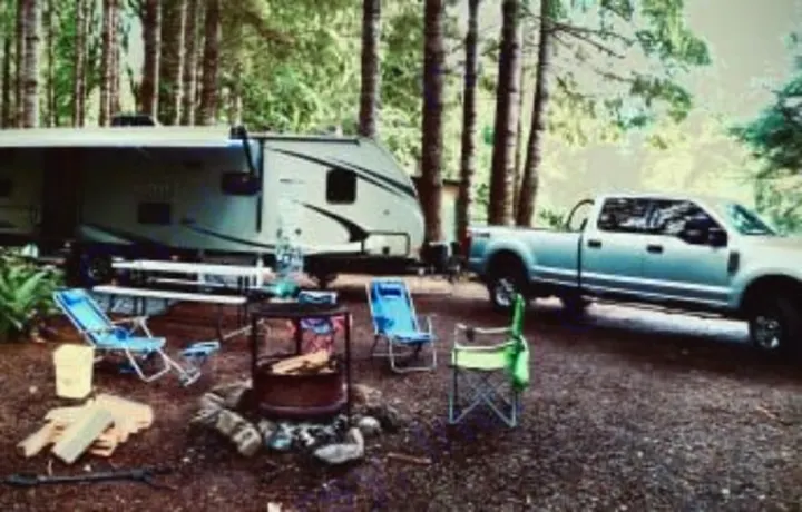 RV rental setup: A travel trailer and truck at a forest campsite with chairs around a fire pit for outdoor camping fun.. Crossroads RV Sunset Trail Grand Reserve 2018