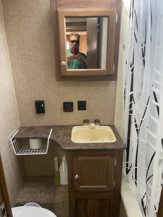 Inside an RV rental: a compact bathroom with vanity, sink, mirror reflecting a man's selfie, shower, and toilet.. KZ Sportsman 2015
