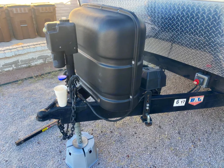 Close-up of an RV rental trailer's front hitch area, showing the propane tank cover, jack stand, and diamond plate siding.. KZ Sportsman 2015