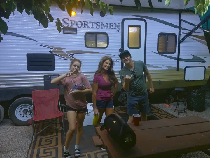 Friends enjoying their RV rental, posing outdoors next to a bright Sportsmen travel trailer at a campsite.. KZ Sportsman 2015