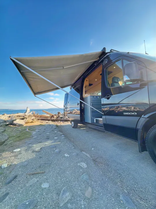 RV rental: Black camper van parked beachside with awning out, door open revealing custom interior. Coastal adventure ready.. Mercedes Sprinter 2007