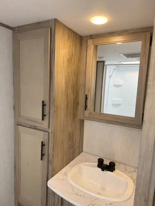 RV rental bathroom features: sink, black faucet, wall-mounted mirror, wood cabinets, and a reflected shower area.. Forest River Cherokee Alpha Wolf 2022