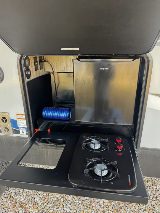 RV rental outdoor kitchen featuring a Hotpoint mini-fridge, Suburban gas stove, and utility connections for camping.. Forest River Cherokee Alpha Wolf 2022