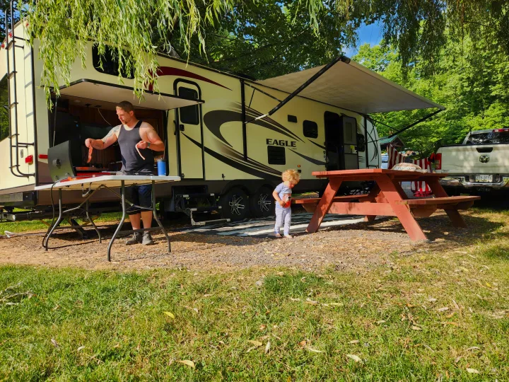 RV rental setup: A man grills food by a large RV, with an awning extended, as a child plays near a picnic table.. Jayco Eagle 2016