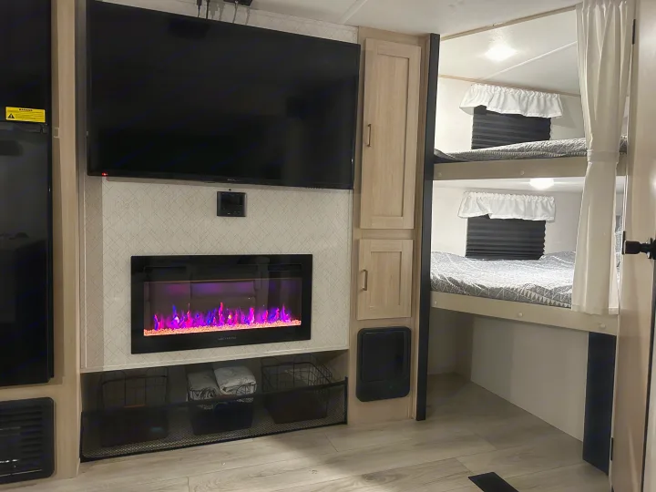 This inviting RV rental interior features a TV over a colorful electric fireplace, with cozy bunk beds and privacy curtains on the right.. Prime Time Avenger 27DBS 2024