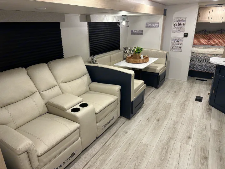 This RV rental interior features comfy recliners, a dinette, and light wood-look flooring, creating a modern and cozy living space.. Prime Time Avenger 27DBS 2024