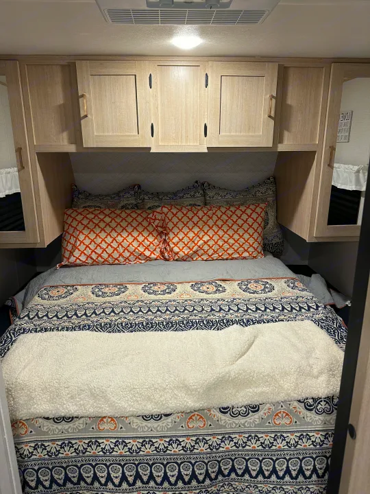Cozy RV rental bedroom featuring patterned bedding, orange pillows, and functional overhead storage cabinets.. Prime Time Avenger 27DBS 2024