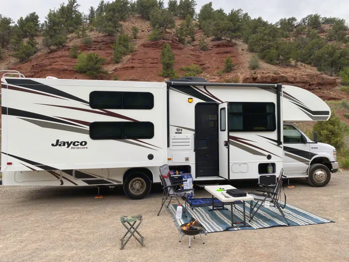 An RV rental with an outdoor campsite setup, including a table, chairs, and grill, against a backdrop of red rock hills.. Jayco Redhawk 2019